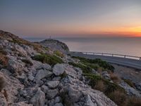 rocks along a road overlooking a large body of water at sunset time with cars driving on the track