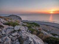 rocks along a road overlooking a large body of water at sunset time with cars driving on the track