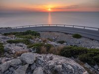 rocks along a road overlooking a large body of water at sunset time with cars driving on the track