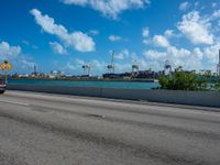 a road going by the ocean with two large ships in the background and blue sky with white clouds