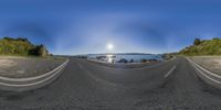 a wide angle view of the ocean and sky and a road leading through it and mountains and a sun in the distance