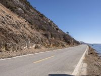 a long empty road next to the ocean in the mountains near some rocks and trees