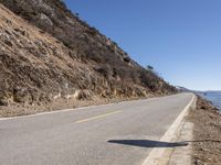 a long empty road next to the ocean in the mountains near some rocks and trees