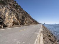 a long empty road next to the ocean in the mountains near some rocks and trees