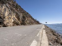 a long empty road next to the ocean in the mountains near some rocks and trees