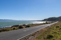 a road by the water and mountains with blue skies in the distance of it is an island on a body of water