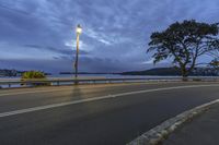 an empty road with street lights by the water with clouds overcasting it, overlooking a city