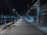 a dark, empty sidewalk lit up at night by street lights and concrete benches under large lamps