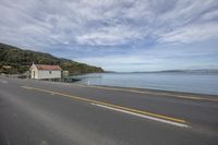 the empty street is along the shore side by the water with a little house on it