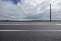 a empty road with a view over the ocean and hills in the background with a cloudy sky