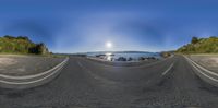 a wide angle view of the ocean and sky and a road leading through it and mountains and a sun in the distance