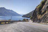 a wide mountain road is flanked by the water and hills with a car driving on the street