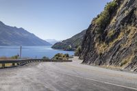 a wide mountain road is flanked by the water and hills with a car driving on the street