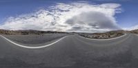 a fish eye view is of a empty road surrounded by rocky hills with cloudy skies