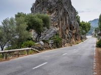 a winding road lined with trees near a mountain range and large rocks in the background