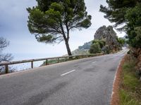 a winding road lined with trees near a mountain range and large rocks in the background