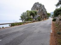 a winding road lined with trees near a mountain range and large rocks in the background