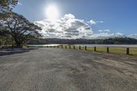 a paved road with wooden posts near water, clouds and trees in background and sun shining