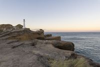 a rocky cliff with a light house in the background as sun sets over the ocean