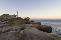 a rocky cliff with a light house in the background as sun sets over the ocean