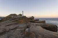 a rocky cliff with a light house in the background as sun sets over the ocean