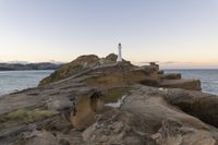 a rocky cliff with a light house in the background as sun sets over the ocean