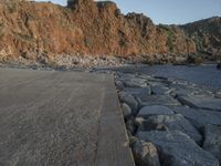 a skateboard parked on a large rocky beach near water at sunrise time with cliff in the background