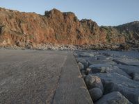 a skateboard parked on a large rocky beach near water at sunrise time with cliff in the background