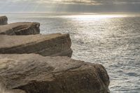 a person stands next to the water looking out to sea, holding a surfboard and standing in front of a rock formation that appears like structure