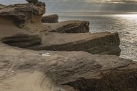 a person stands next to the water looking out to sea, holding a surfboard and standing in front of a rock formation that appears like structure