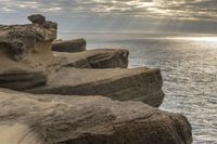 a person stands next to the water looking out to sea, holding a surfboard and standing in front of a rock formation that appears like structure