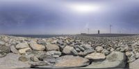 several large rocks piled up by the ocean in front of a cloudy sky, and a small lighthouse behind them