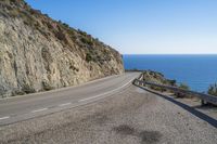 a narrow paved road on a cliff overlooking the ocean with cars parked in front of it