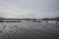 a wooden pier next to a river on a cloudy day near trees and buildings and mountains