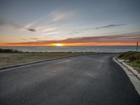 the sun is setting on the horizon with an empty road in front of it and ocean in back