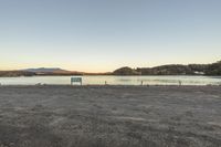 a bench sitting in the middle of a dirt lot next to water and hills in the background