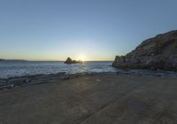 a person sitting on a bench in front of the water at sunset near some rocks