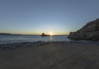 a person sitting on a bench in front of the water at sunset near some rocks