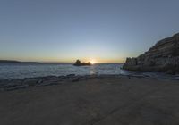 a person sitting on a bench in front of the water at sunset near some rocks