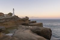 the lighthouse is perched on top of some rocks next to the water and cliffs at sunset