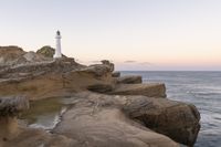 the lighthouse is perched on top of some rocks next to the water and cliffs at sunset