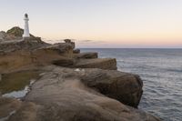 the lighthouse is perched on top of some rocks next to the water and cliffs at sunset