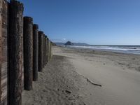 a line of old wooden posts along a sandy beach shoreline to sea water and mountains in the distance