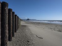 a line of old wooden posts along a sandy beach shoreline to sea water and mountains in the distance