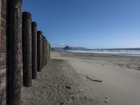 a line of old wooden posts along a sandy beach shoreline to sea water and mountains in the distance