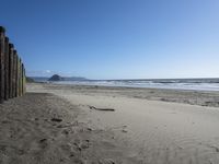 a line of old wooden posts along a sandy beach shoreline to sea water and mountains in the distance