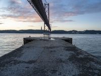 the sun sets over the water and the bridge is high above it, with an object in the foreground