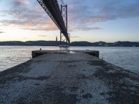 the sun sets over the water and the bridge is high above it, with an object in the foreground