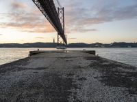 the sun sets over the water and the bridge is high above it, with an object in the foreground