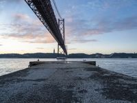 the sun sets over the water and the bridge is high above it, with an object in the foreground